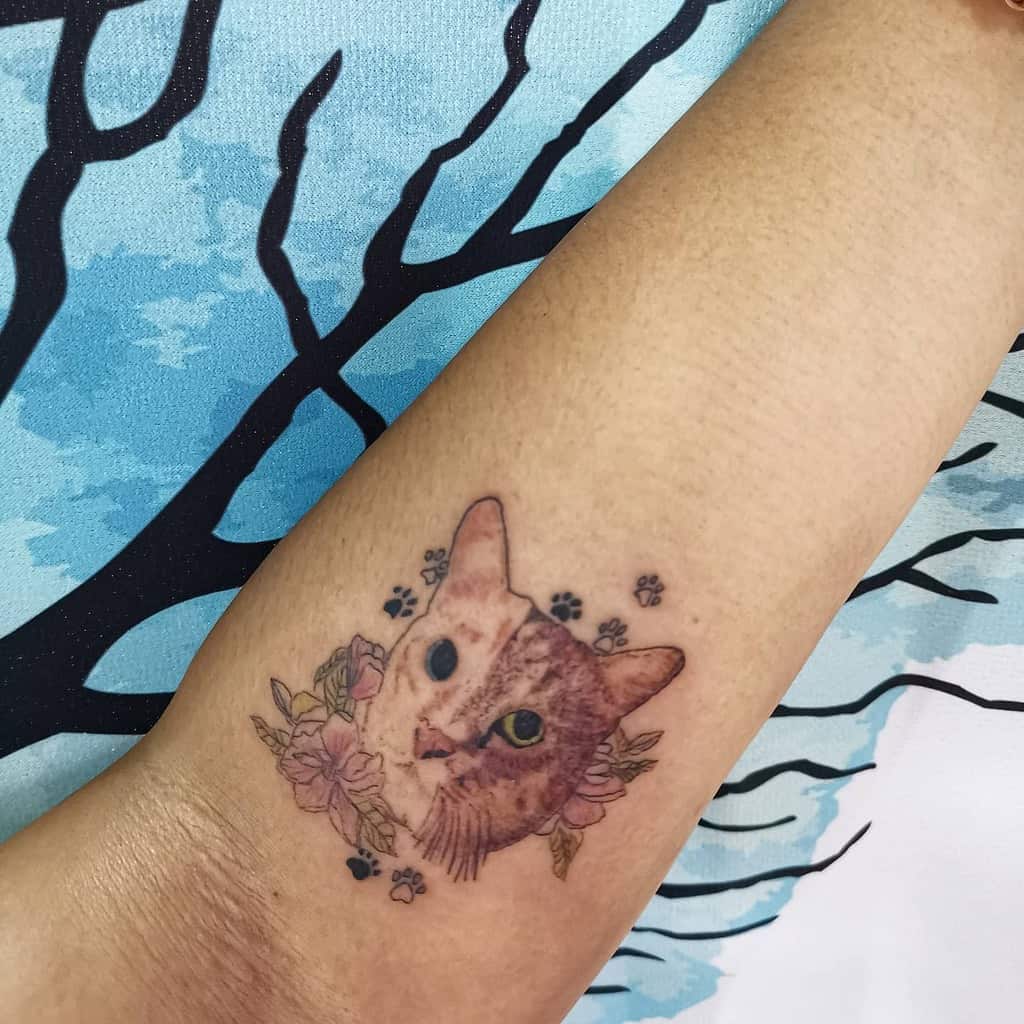 Forearm tattoo of a detailed cat portrait surrounded by pink flowers and tiny paw prints, designed with soft colors