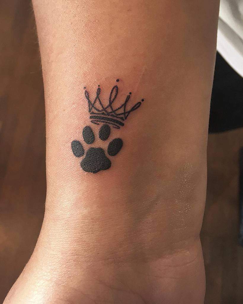 Tattoo of a sleek black cat paw print with a delicate crown above it on a person's inner forearm