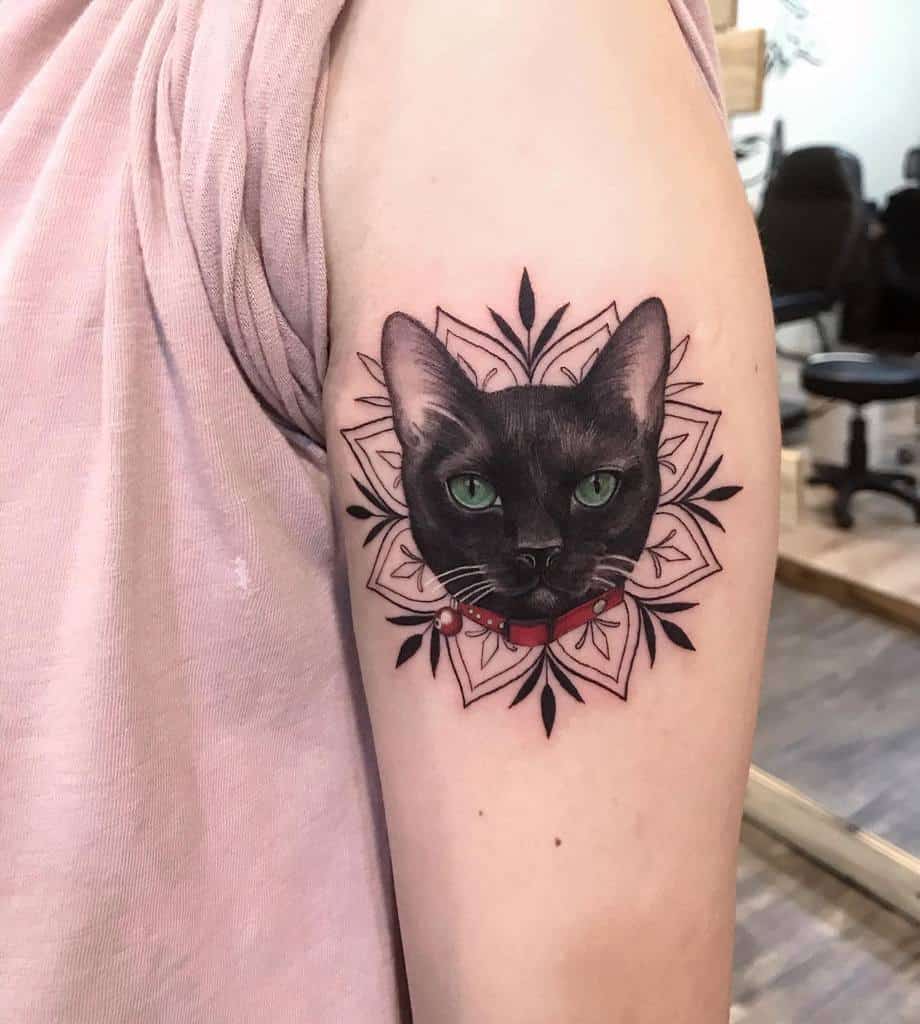 Tattoo of a black cat's face with green eyes, framed by a geometric floral pattern on an arm