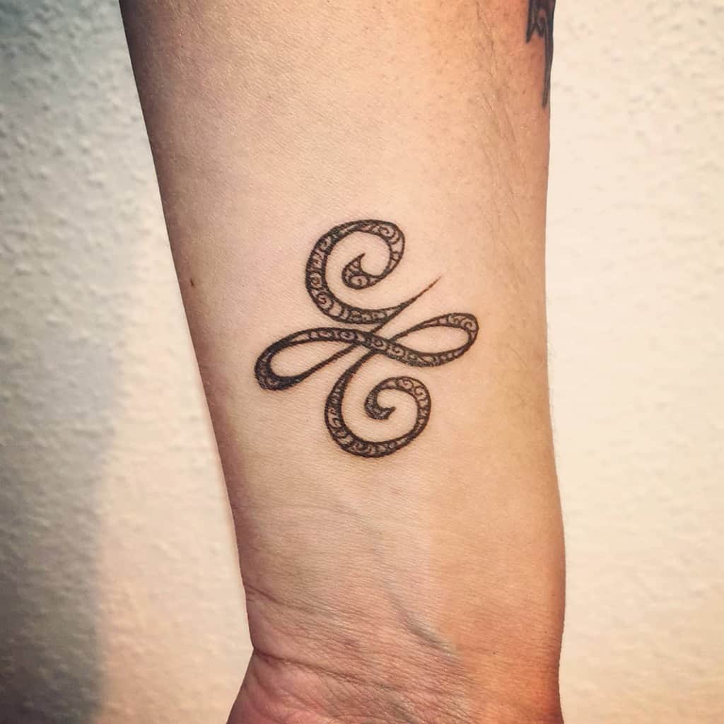 Ornate scrollwork tattoo resembling an ampersand symbol on forearm, with intricate swirling patterns
