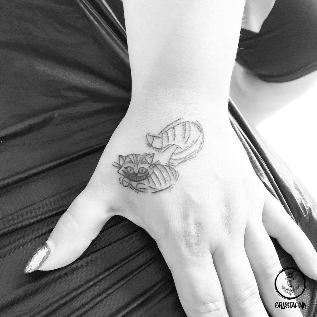 A small, minimalist Cheshire Cat tattoo on the hand, featuring its signature grin and curled tail