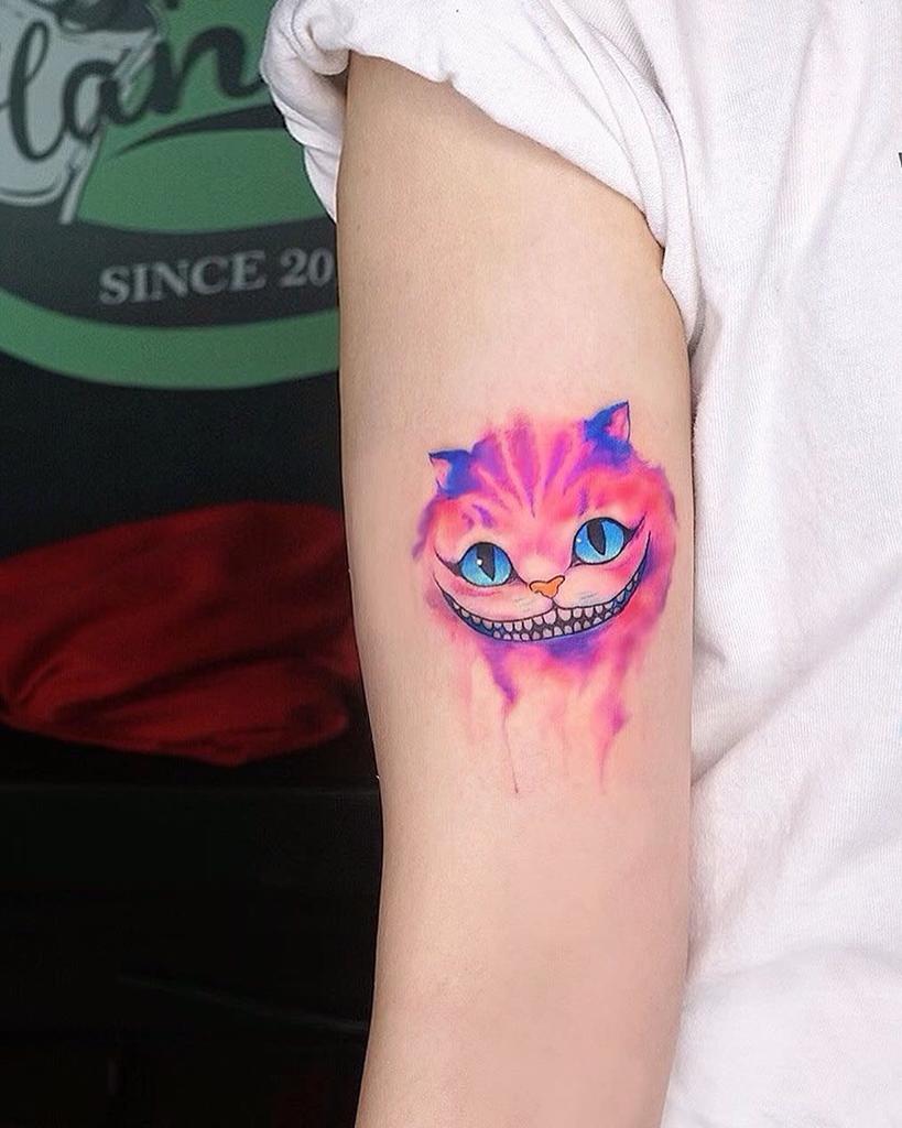 A vibrant watercolor-style Cheshire Cat tattoo on the arm, featuring bright blue eyes and a wide grin