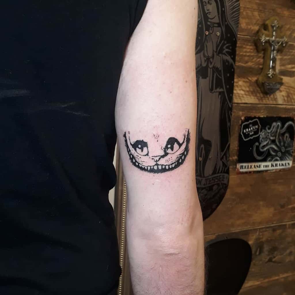 A minimalist black ink tattoo of the Cheshire Cat’s eyes and wide grin on the upper arm, capturing its eerie smile