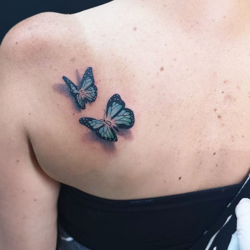 Two teal and black butterfly tattoos on the upper back, featuring soft shading and delicate wing details for a vibrant look