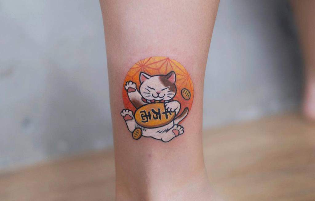 Vibrant tattoo of a lucky cat holding a gold coin with an orange circular background, symbolizing prosperity and good fortune
