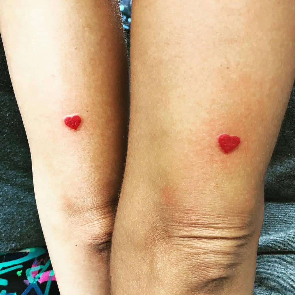 Two arms with small red heart tattoos near the elbows