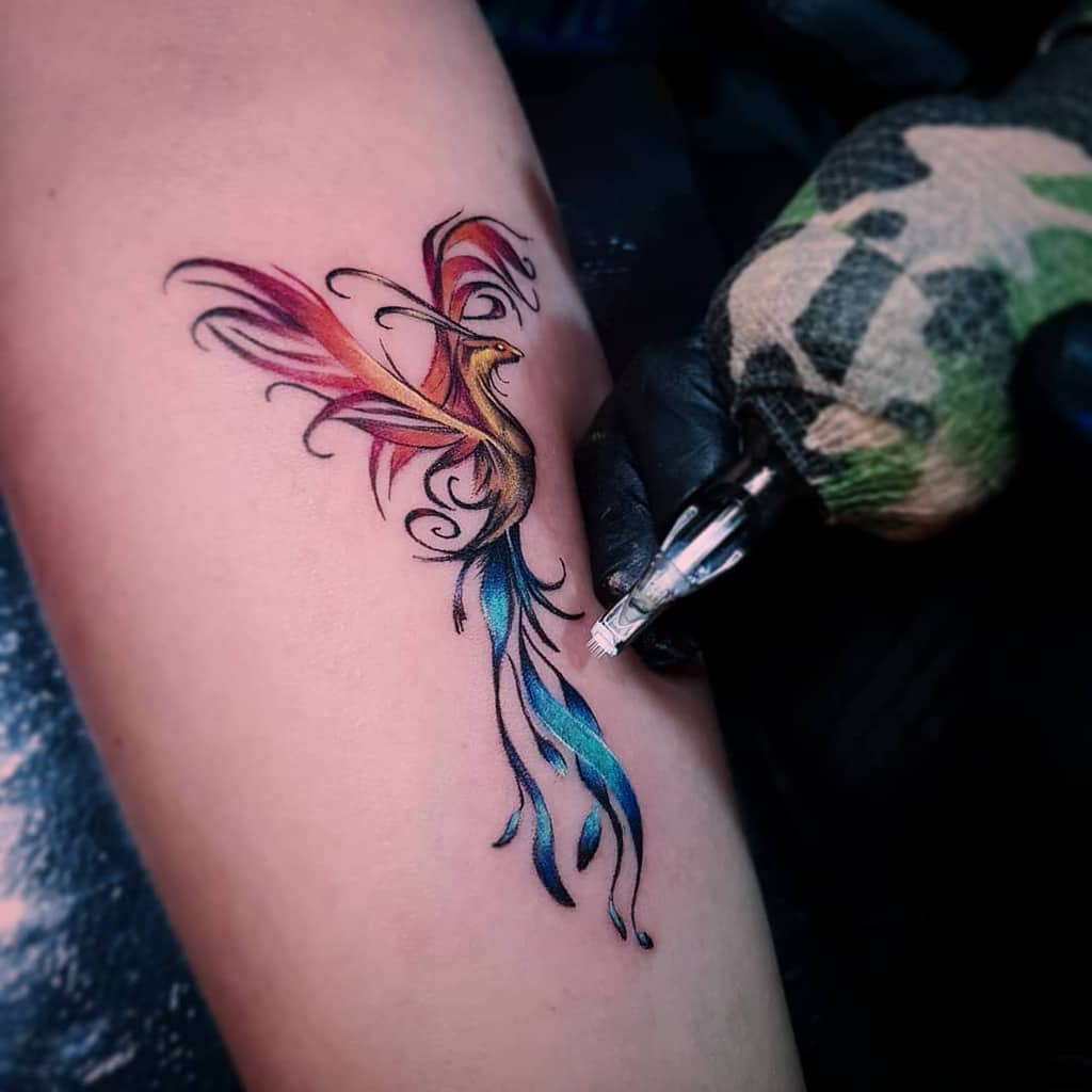 A vibrant, colorful phoenix tattoo with fiery red and yellow wings and flowing blue and green tail feathers, in the process of being inked