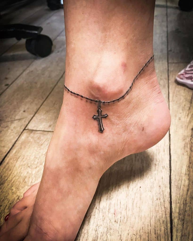 Ankle tattoo of a cross hanging from a delicate chain design, resembling a rosary for a meaningful look