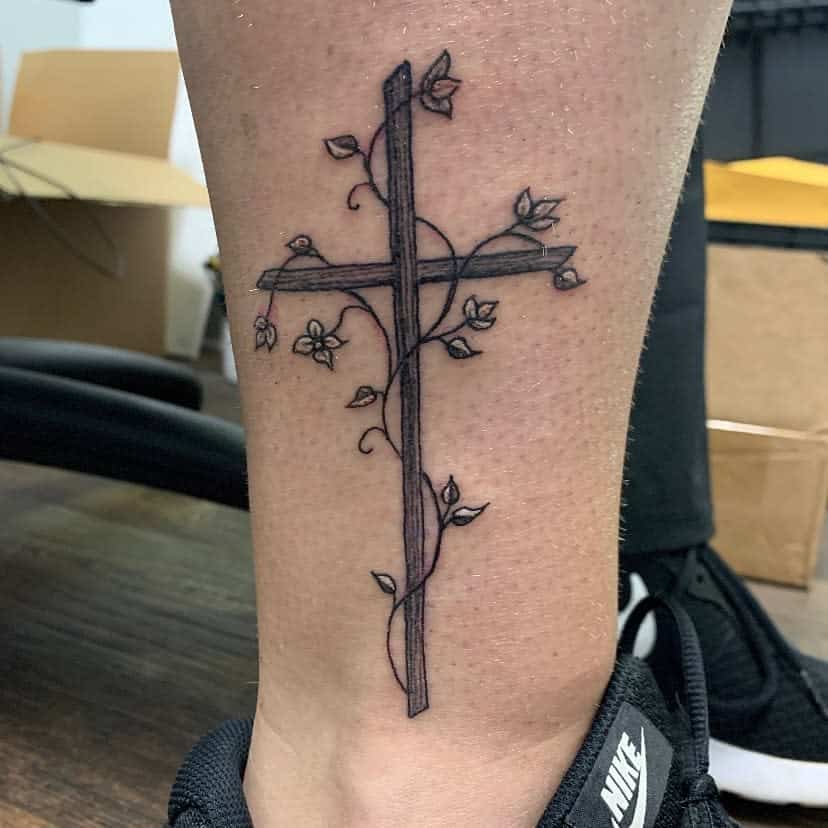 Tattoo of a wooden cross with vines and flowers wrapped around it on a  leg