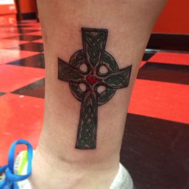 Tattoo of a Celtic cross with green patterns and a red heart at the center on a lower leg