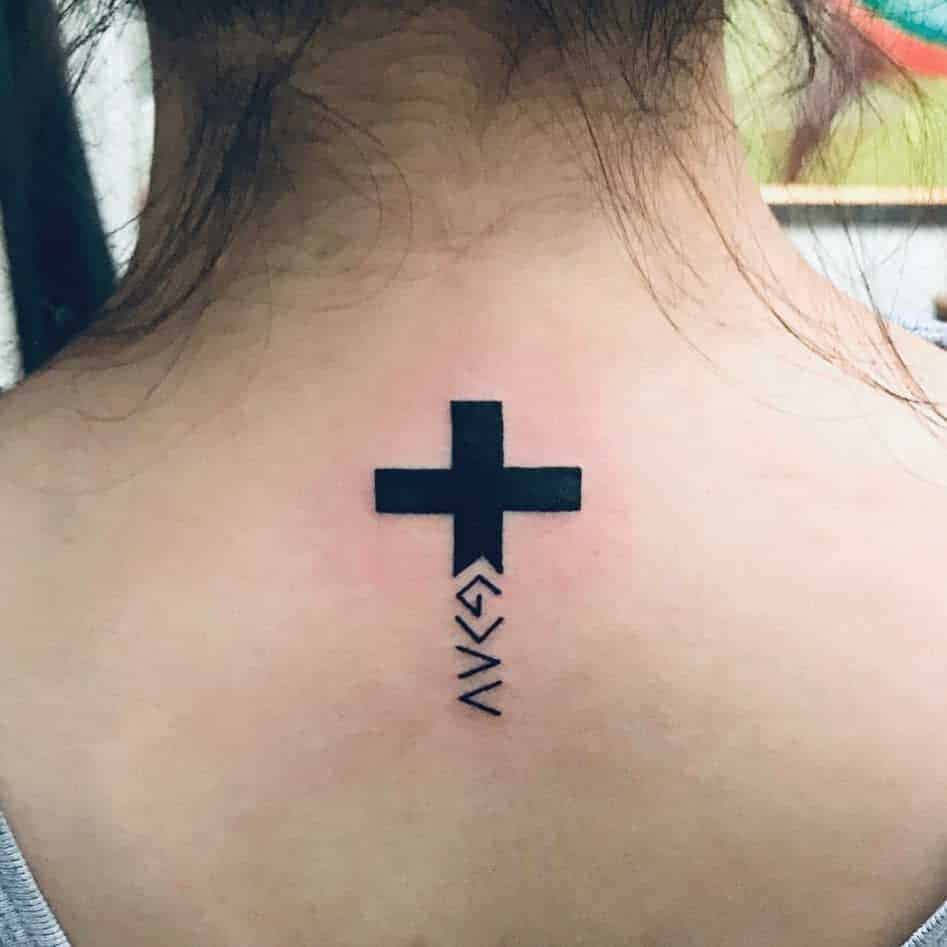 Tattoo of a black cross on a person's upper back with symbols or letters underneath