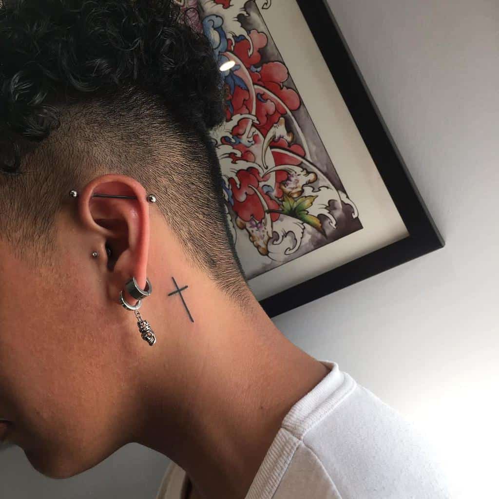 Simple black cross tattoo behind the ear, blending subtle elegance with a spiritual touch