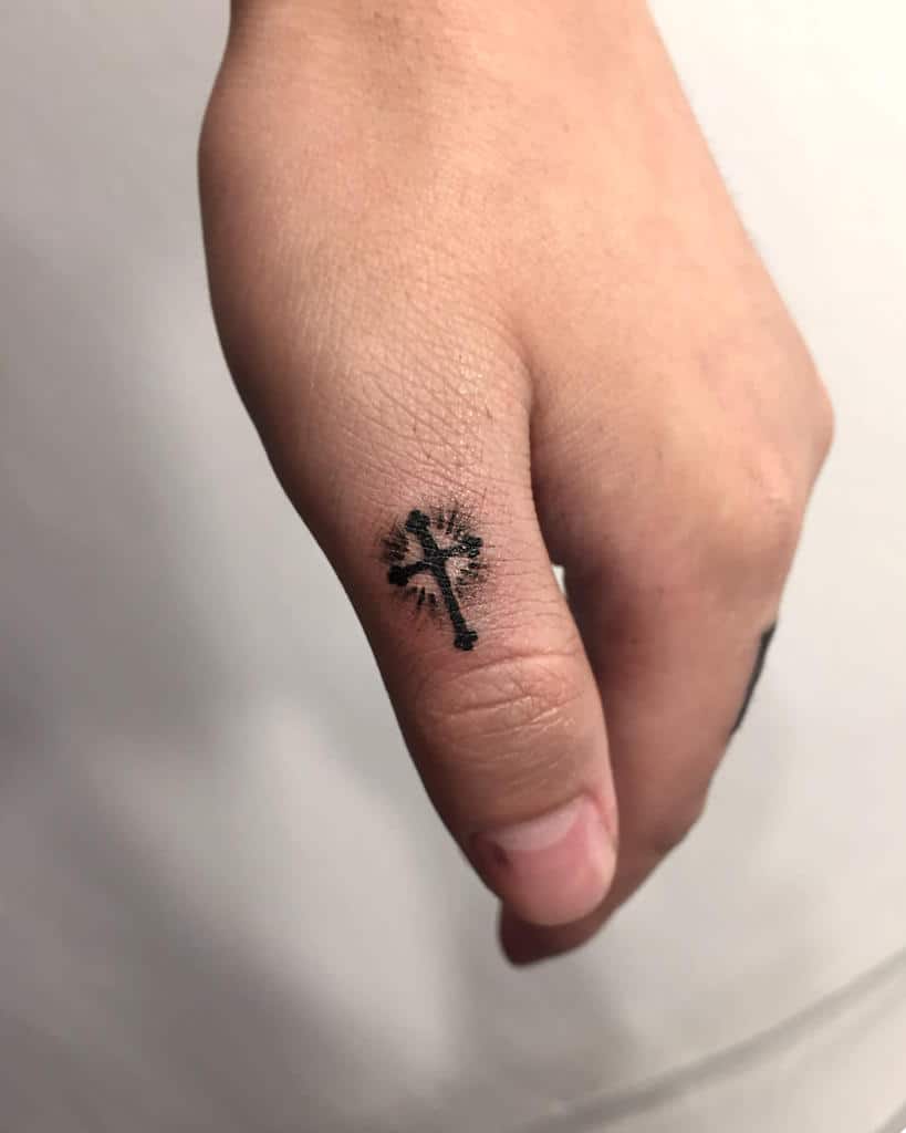 Tiny cross tattoo on the side of the finger, surrounded by radiating lines for a simple yet radiant design