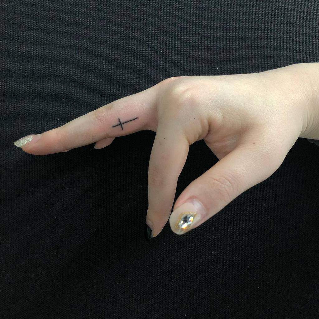 A hand with a small black cross tattoo on the middle finger