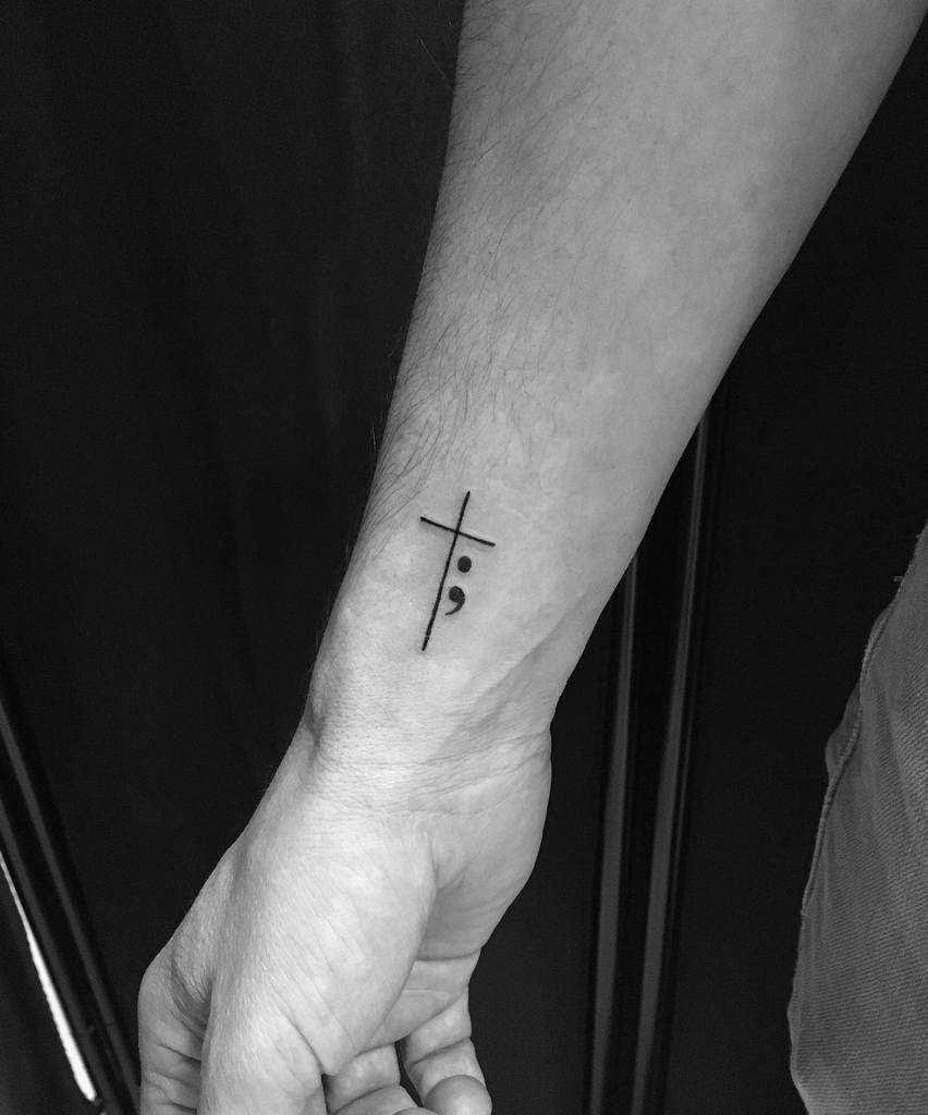 Wrist with a tattoo of a cross and semicolon symbol on the inner forearm in black ink