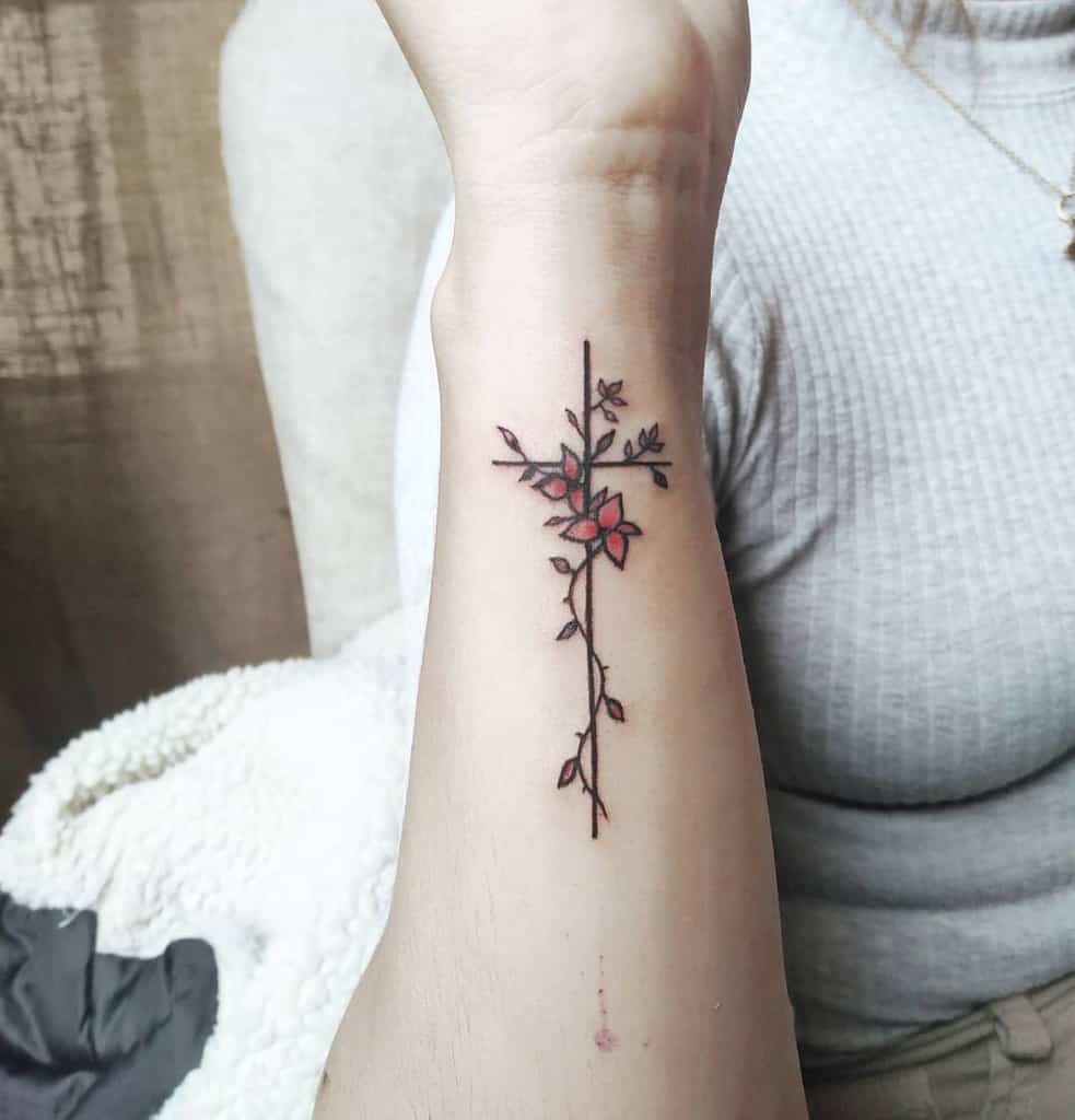 A cross tattoo with floral vines and red flower details on a forearm