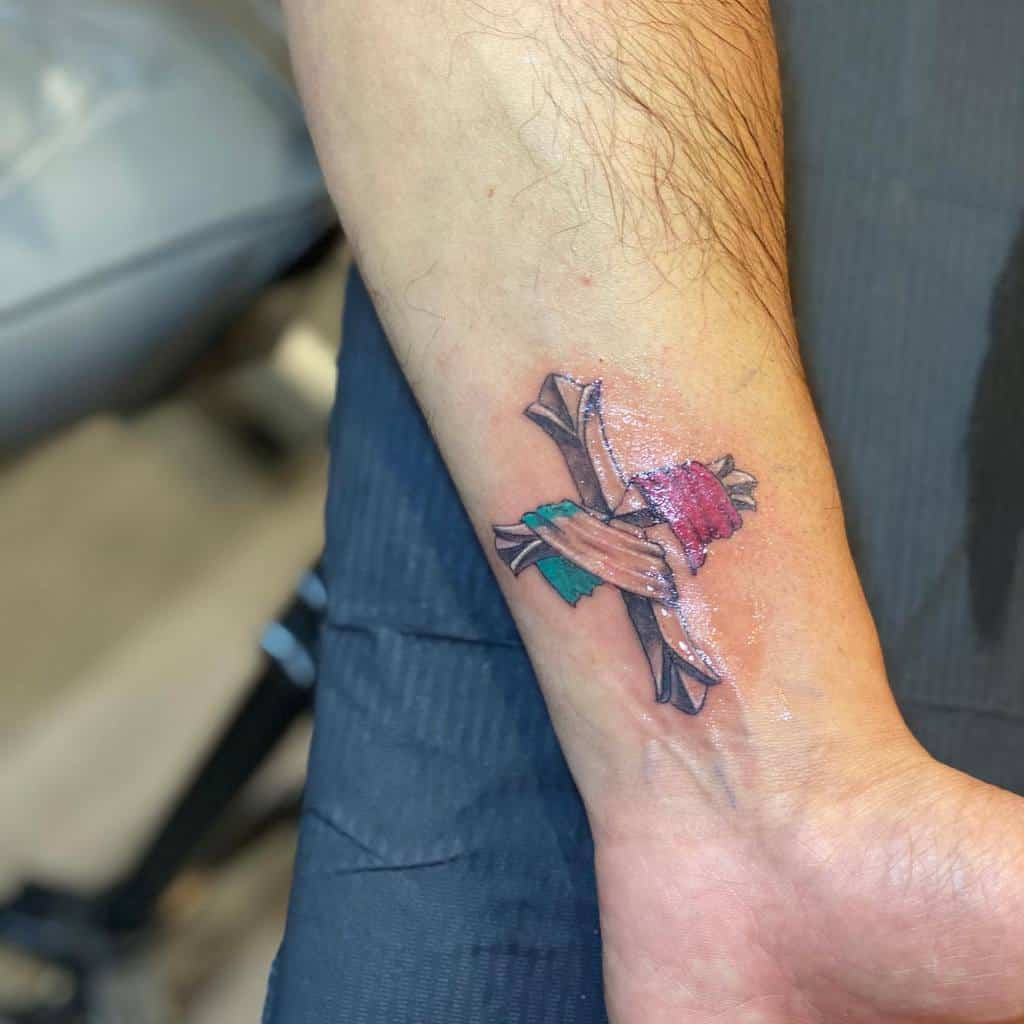 Tattoo of a wooden crucifix wrapped in a red, green, and white cloth on a wrist