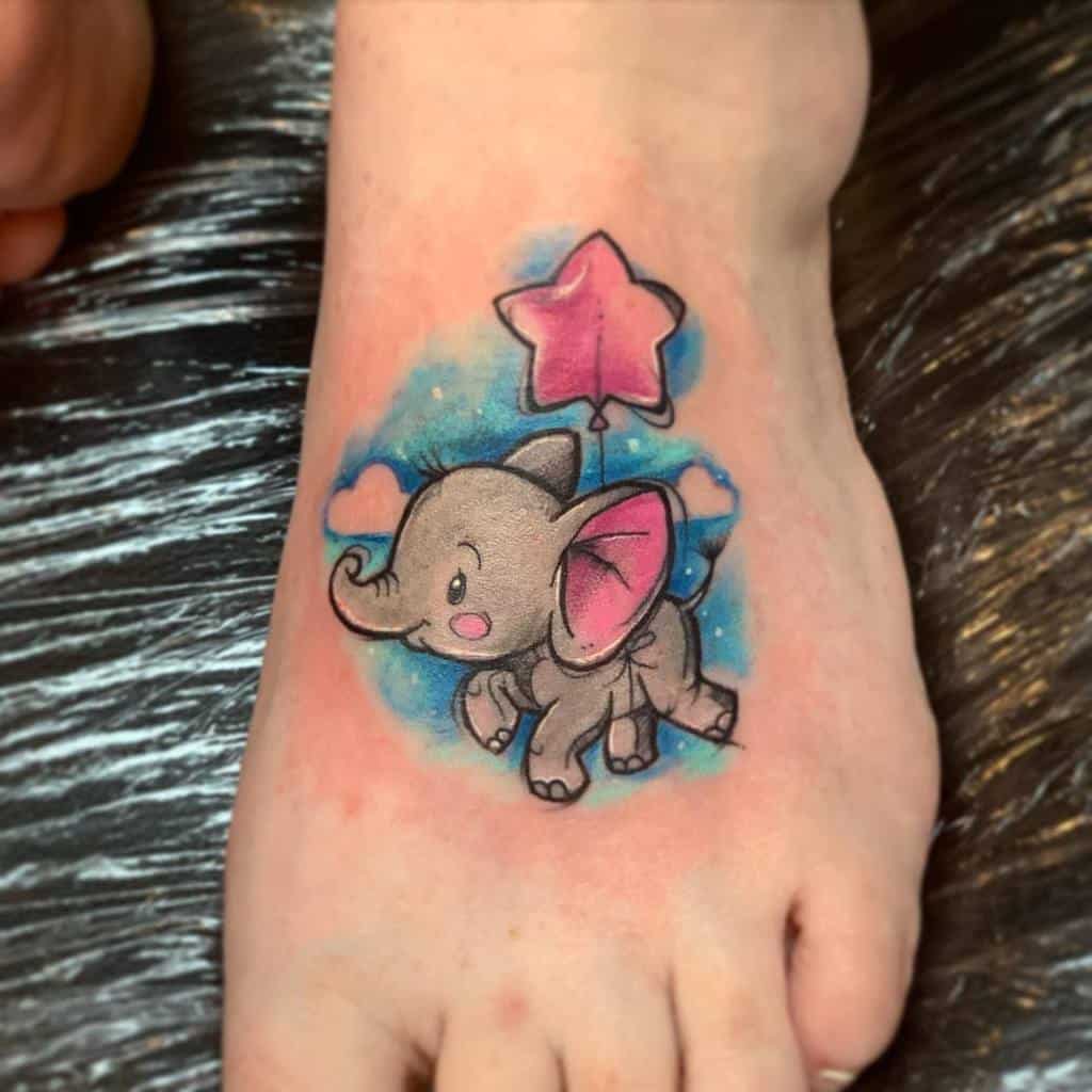 A charming tattoo of Dumbo the elephant, flying with a star-shaped balloon, surrounded by soft blue and pink watercolor effects on the foot