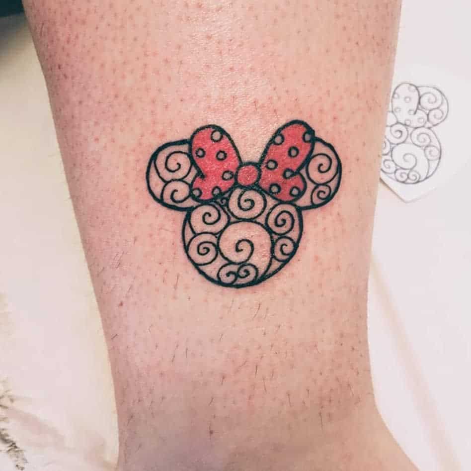 A delicate tattoo of Minnie Mouse's iconic ears and red polka dot bow, outlined with swirling patterns, placed on the ankle