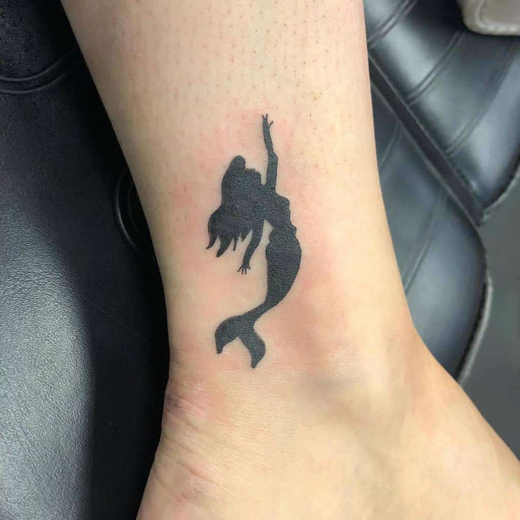 A sleek silhouette tattoo of a mermaid gracefully swimming, inked in black on the ankle