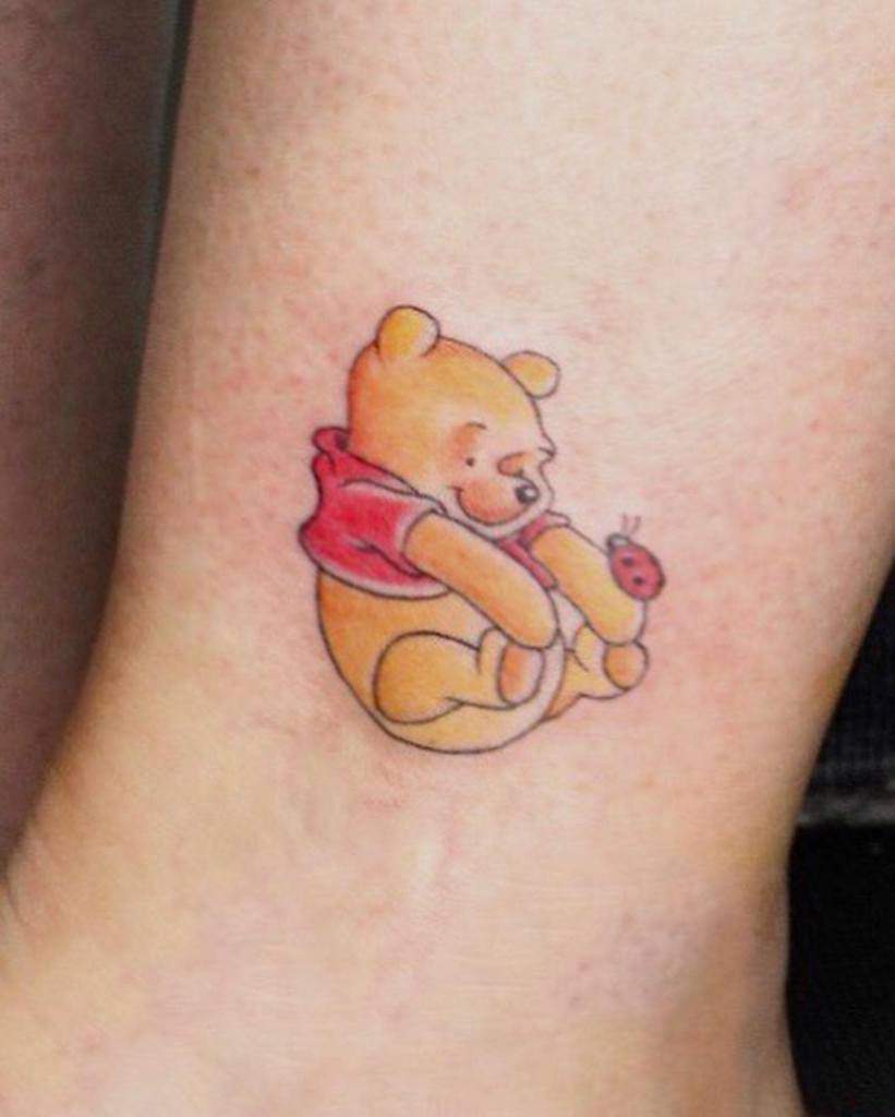 A colorful tattoo of Winnie the Pooh sitting and playing with a ladybug, designed on the ankle in a charming, cartoonish style