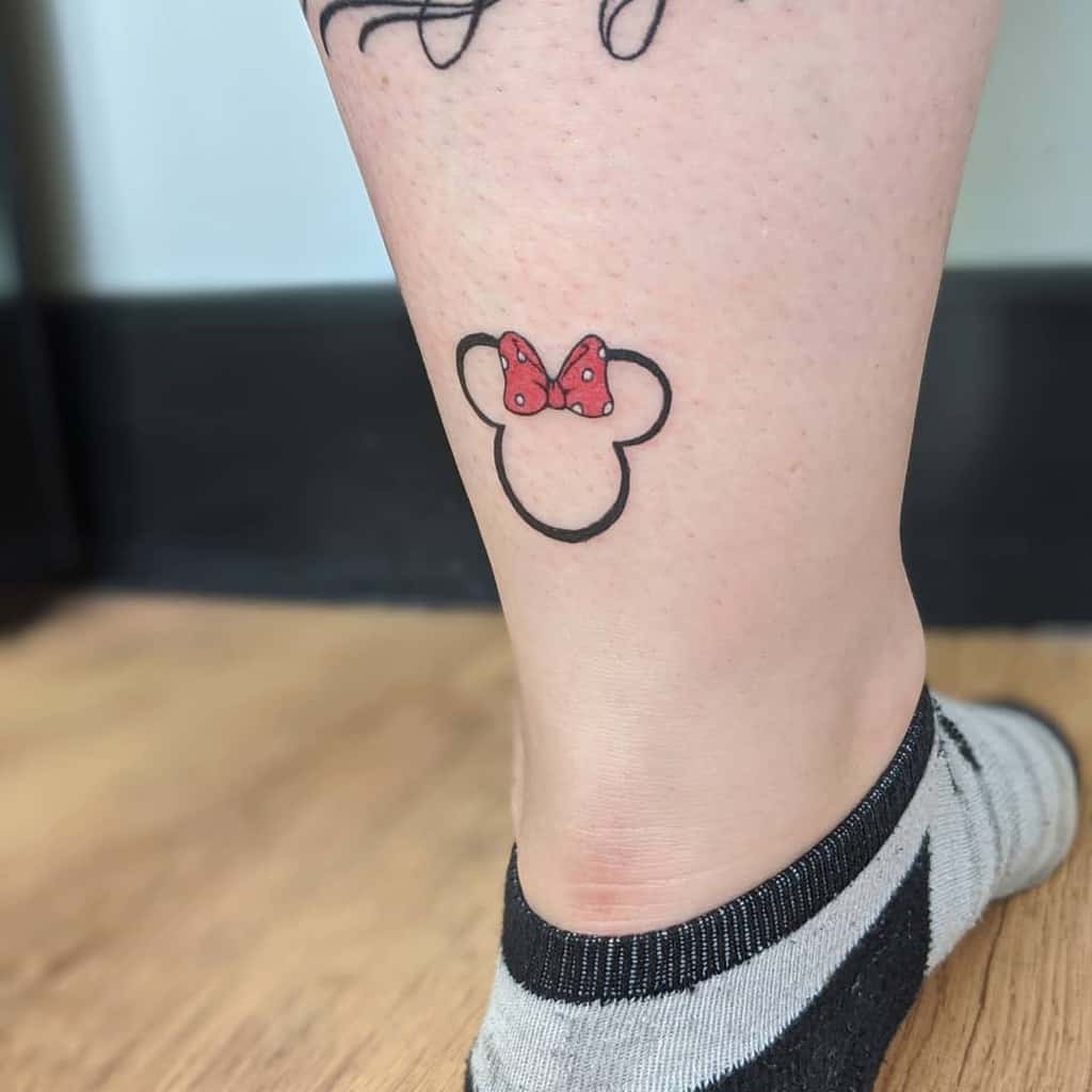 A minimalist tattoo of Minnie Mouse's ears outlined with a red polka dot bow, delicately placed on the ankle