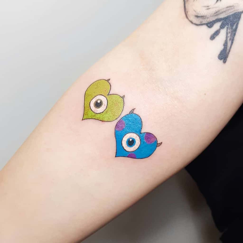 A playful tattoo featuring heart-shaped designs with eyes, inspired by Mike and Sulley from Monsters, Inc., with vibrant colors and whimsical details on the forearm