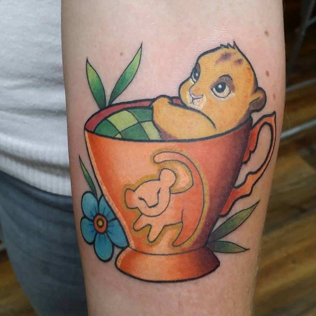 This cute tattoo features Simba as a cub nestled in a teacup, surrounded by playful floral elements, evoking a sense of warmth and innocence