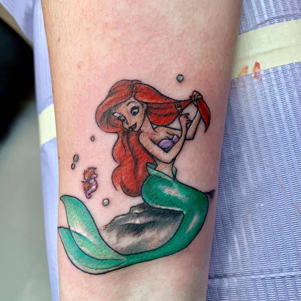 A playful tattoo of Ariel from The Little Mermaid with her signature red hair, green tail, and a small shrimp by her side