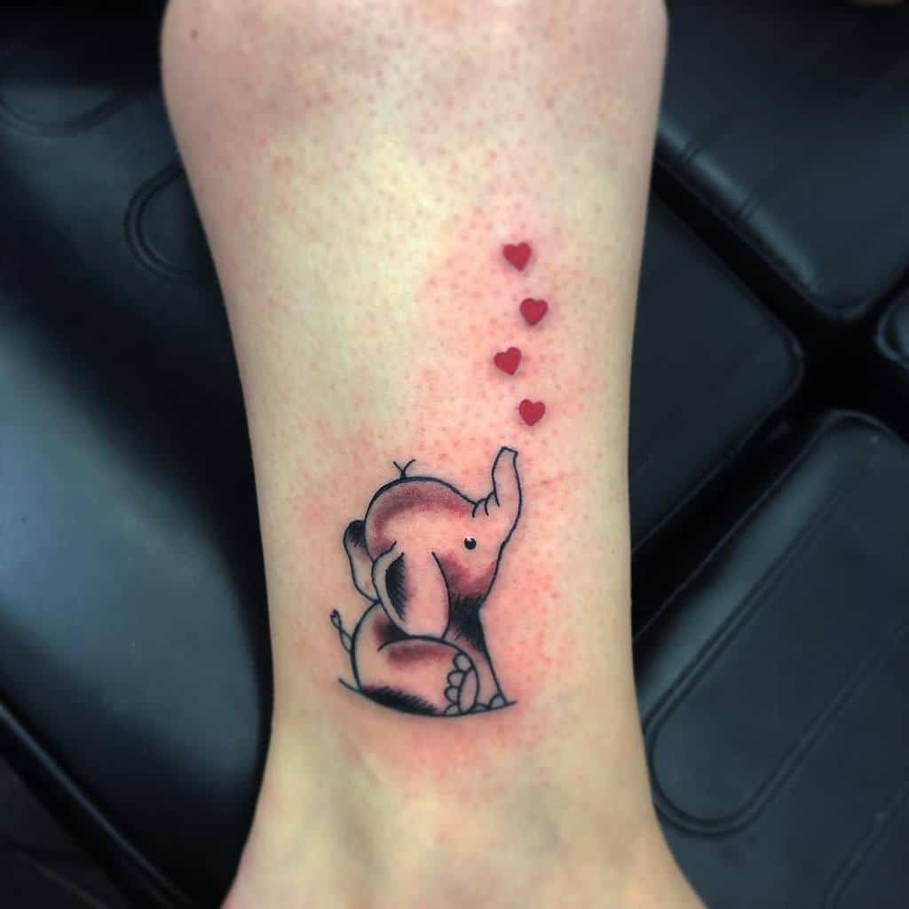 Tattoo of a small elephant with its trunk up, blowing four red hearts, on an ankle