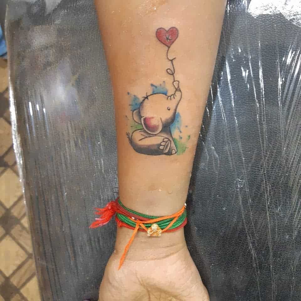 Tattoo of a cartoon elephant holding a heart-shaped balloon on a forearm with colorful thread bracelets