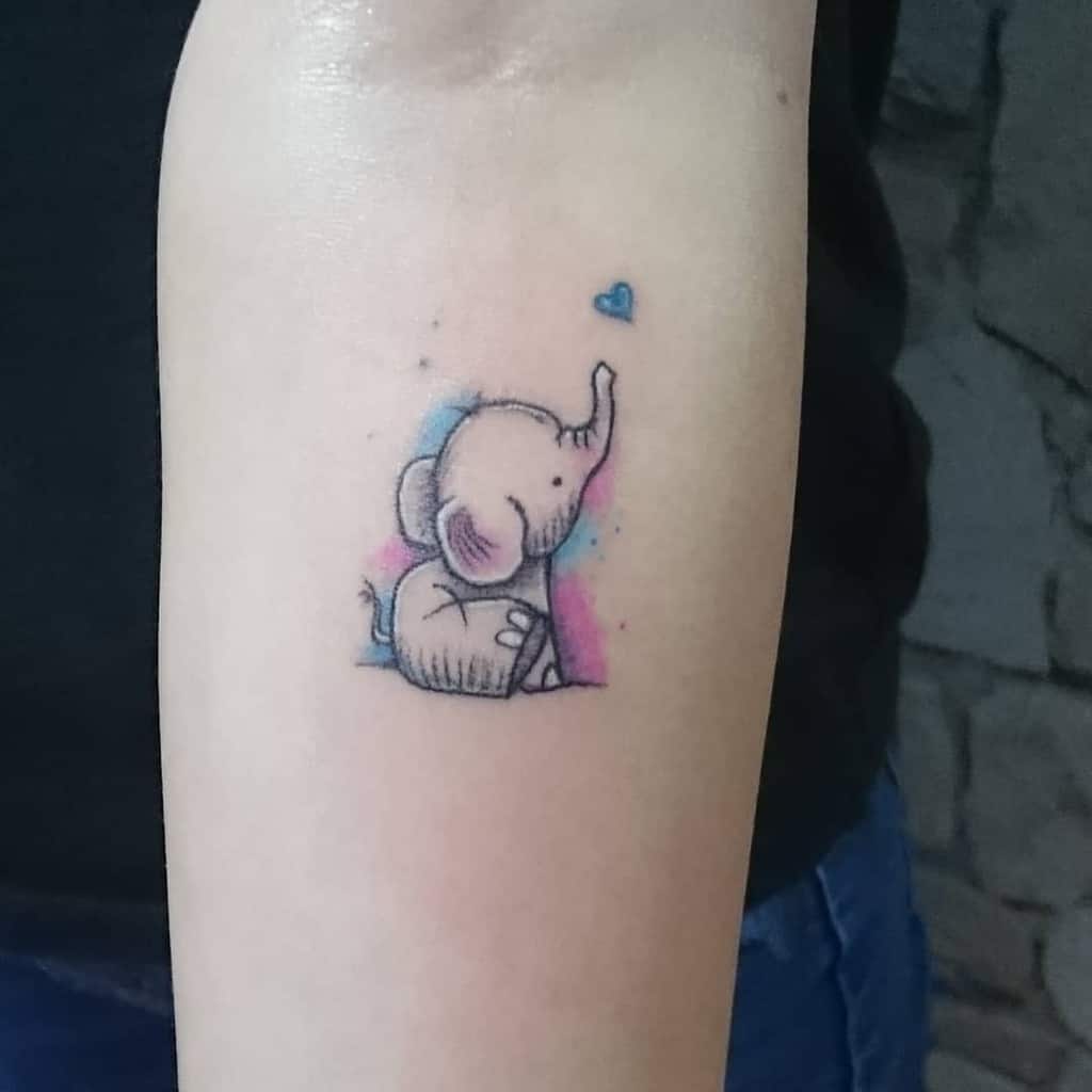 Cute elephant tattoo on arm with a small blue heart above it, featuring soft pink and blue shading