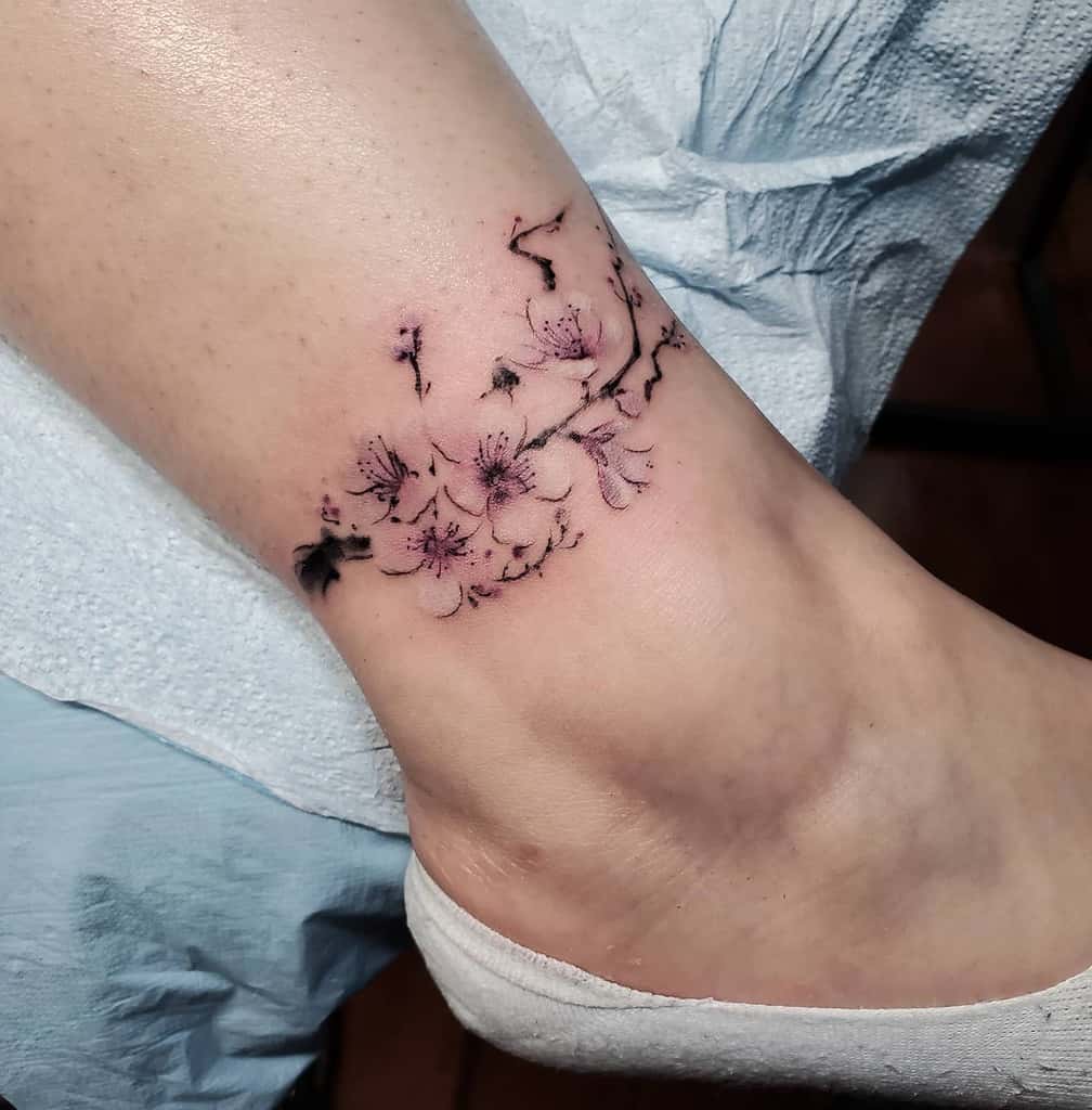 Delicate cherry blossom tattoo encircling an ankle, with light pink petals and subtle black branches
