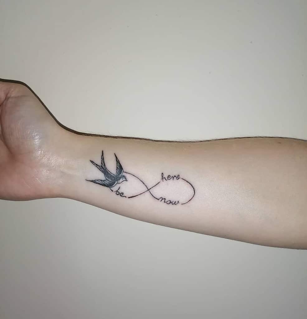 Tattoo of a bird and infinity symbol with "be here now" on a woman's forearm