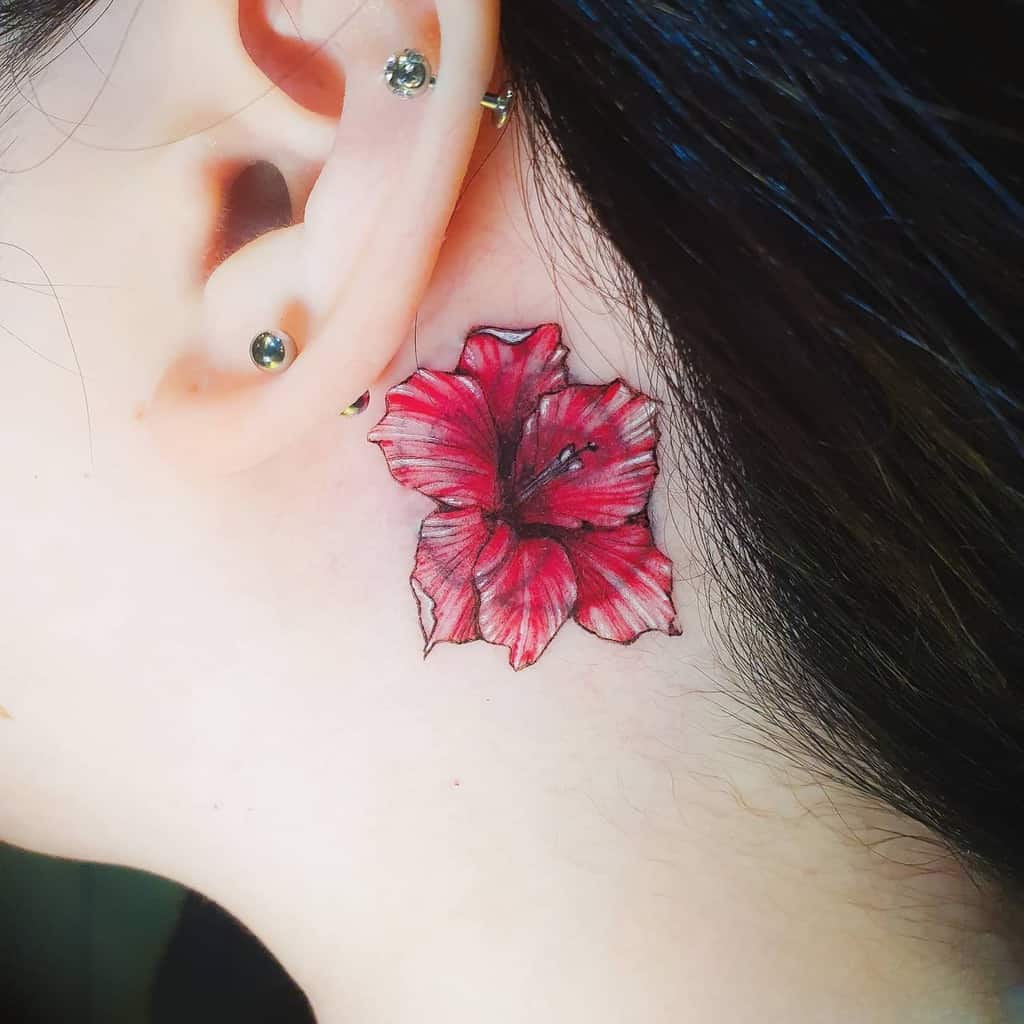Close-up of a gladiolus tattoo, elegantly inked in red, nestled behind the ear of a person with multiple ear piercings