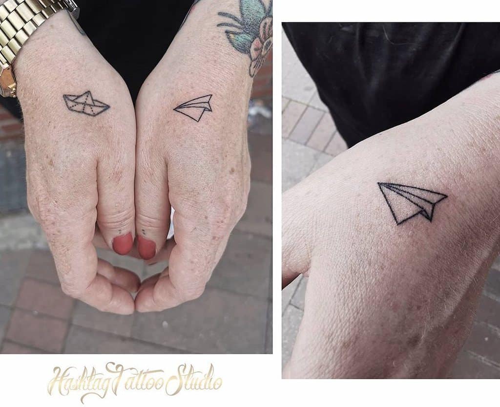 Two hands with tattoos: a paper boat on one hand and a paper airplane on the other, both simple line drawings