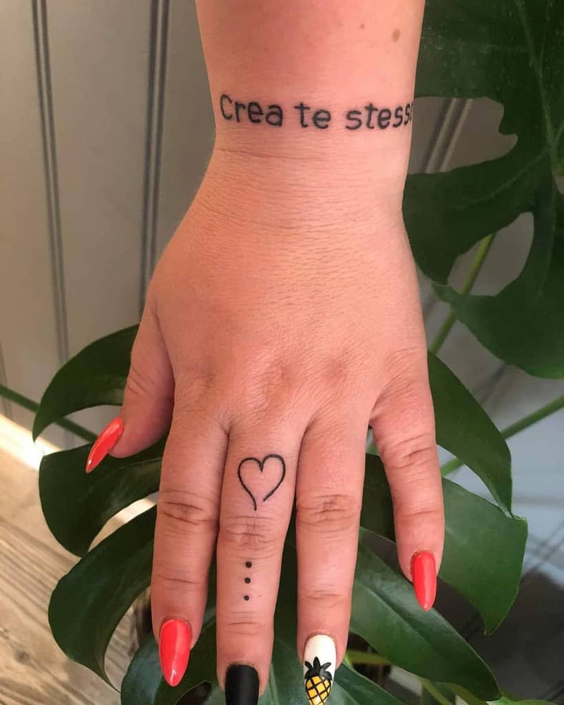 Delicate finger tattoo featuring a minimalist heart and three dots, paired with the phrase 'Crea te stesso' on the wrist