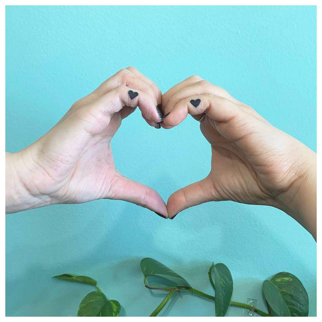 Two hands forming a heart shape with small heart tattoos on thumbs