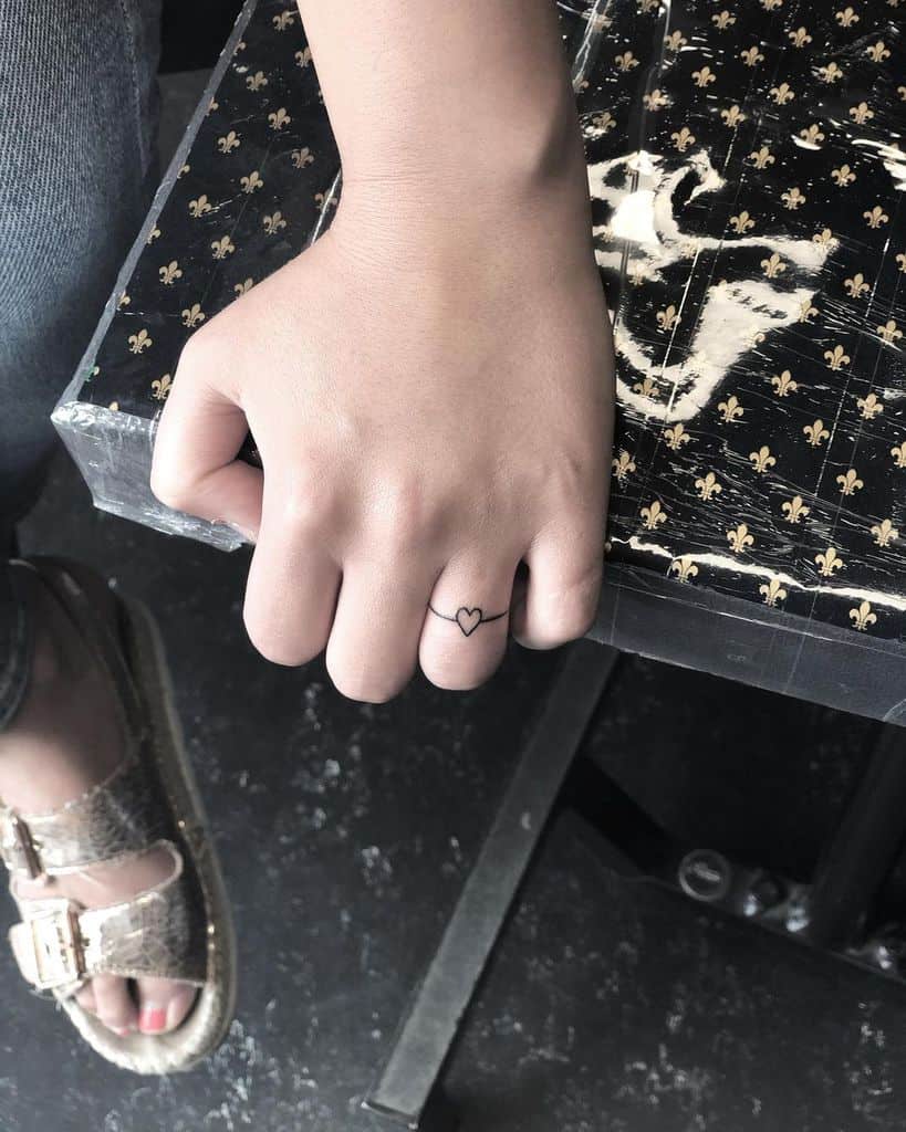 Minimalist finger tattoo featuring a small heart outline connected by a thin band around the finger