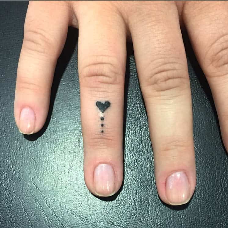Finger with small black heart tattoo on the middle joint followed by three dots