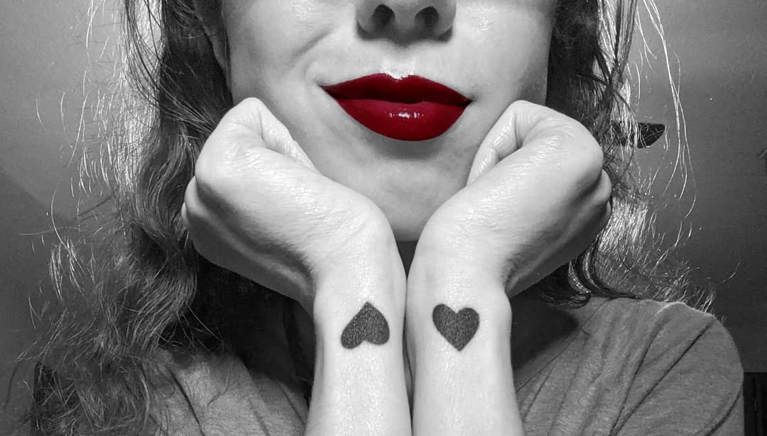 Matching black heart tattoos on wrists, paired with vibrant red lips highlighted in a black-and-white photo