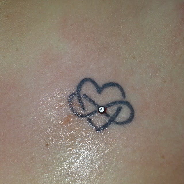 A tattoo of an infinity heart with a small gem in the center graces the skin