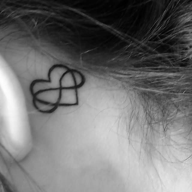 Delicately placed behind the ear, a black tattoo intertwines an infinity symbol with a heart, blending eternal love and boundless connection