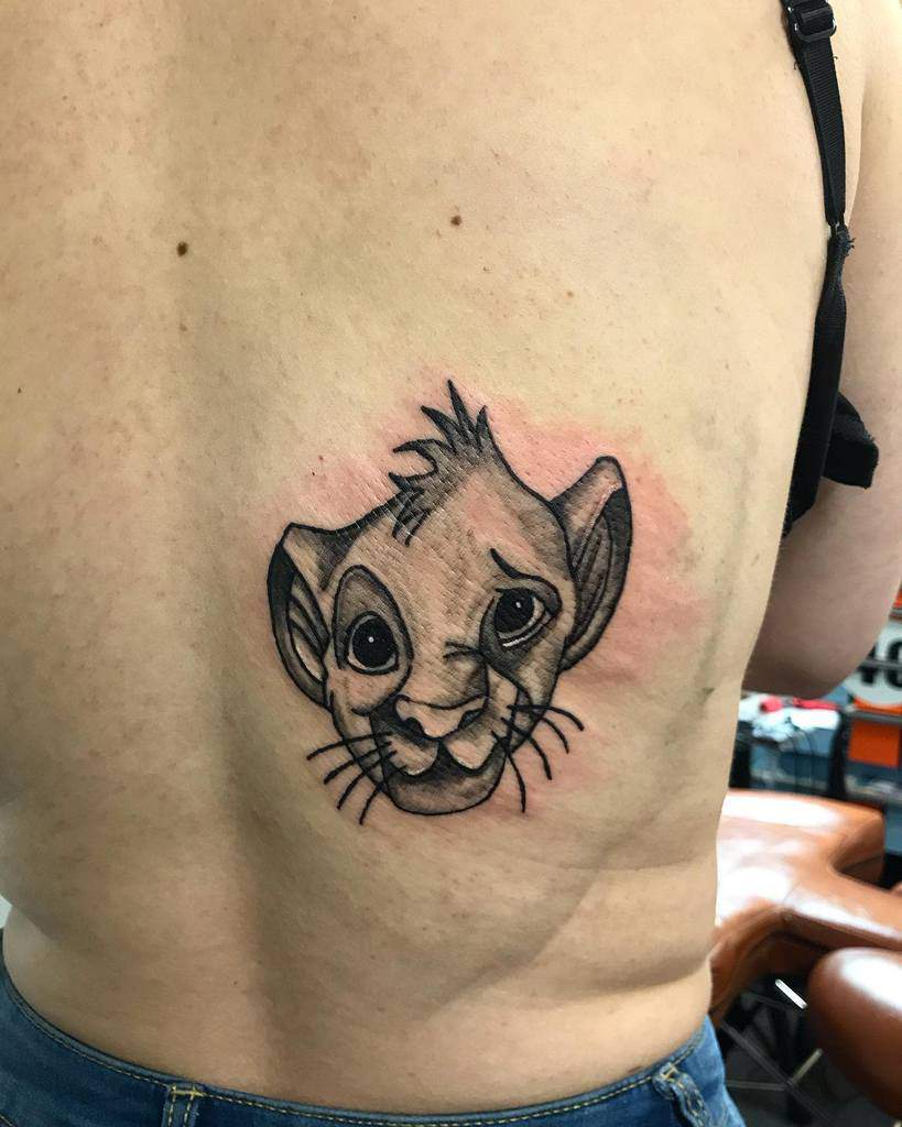 A small detailed black and gray tattoo of a young Simba from The Lion King, placed on the back