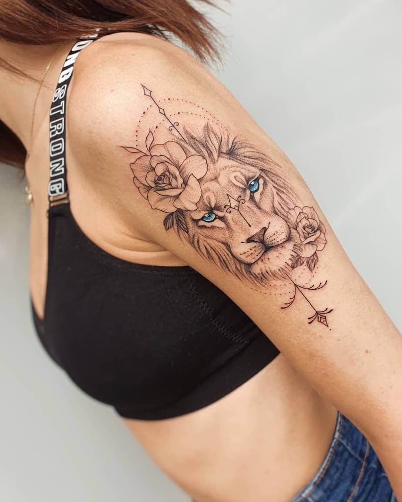 A detailed tattoo on the upper arm featuring a lion's face with blue eyes, surrounded by roses, a geometric arrow, and decorative dots