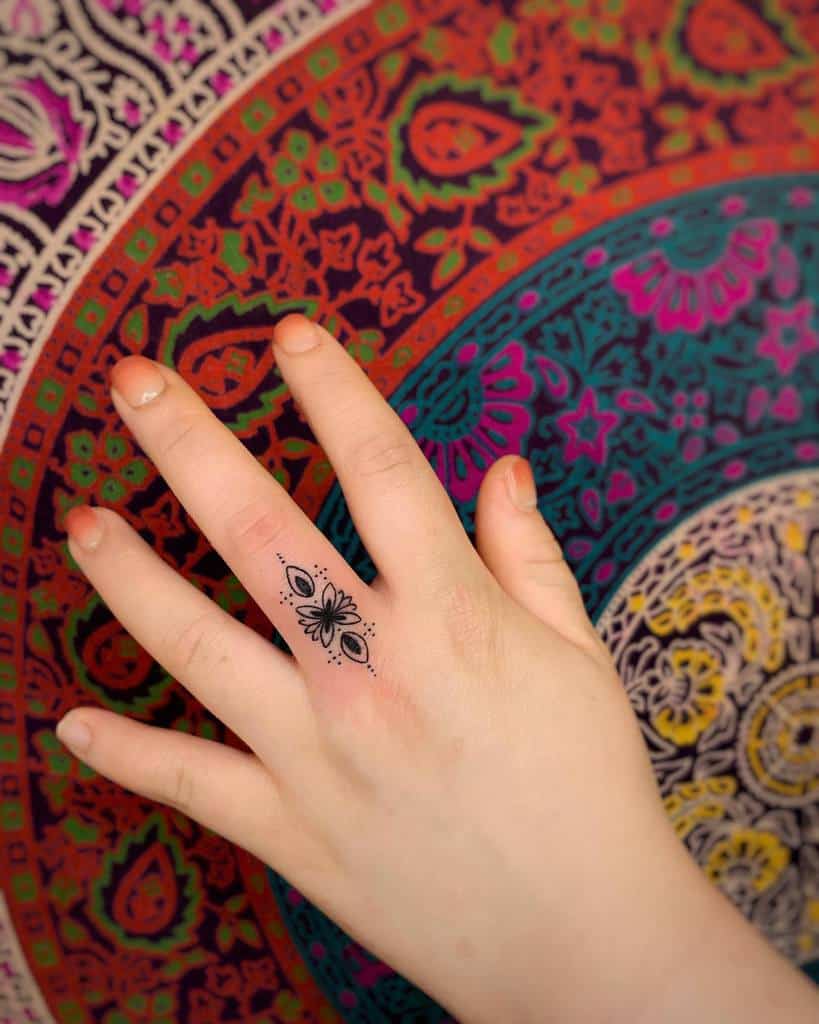 Delicate floral finger tattoo with symmetrical patterns and dot accents, set against a vibrant mandala background