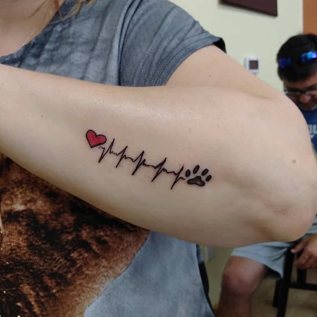 A forearm tattoo of a heartbeat line with a red heart and a paw print at the end