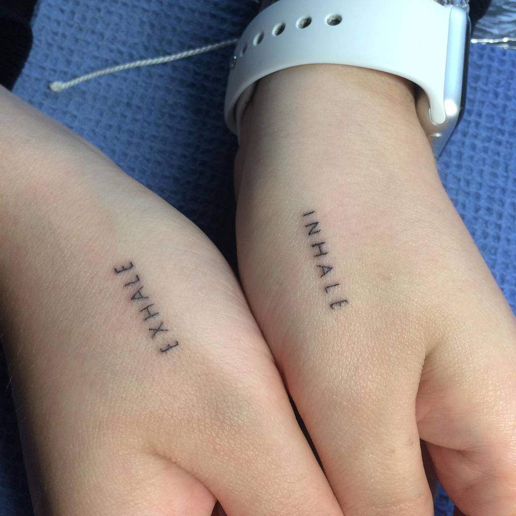 Two wrists with small tattoos: "INHALE" on the right wrist and "EXHALE" on the left wrist