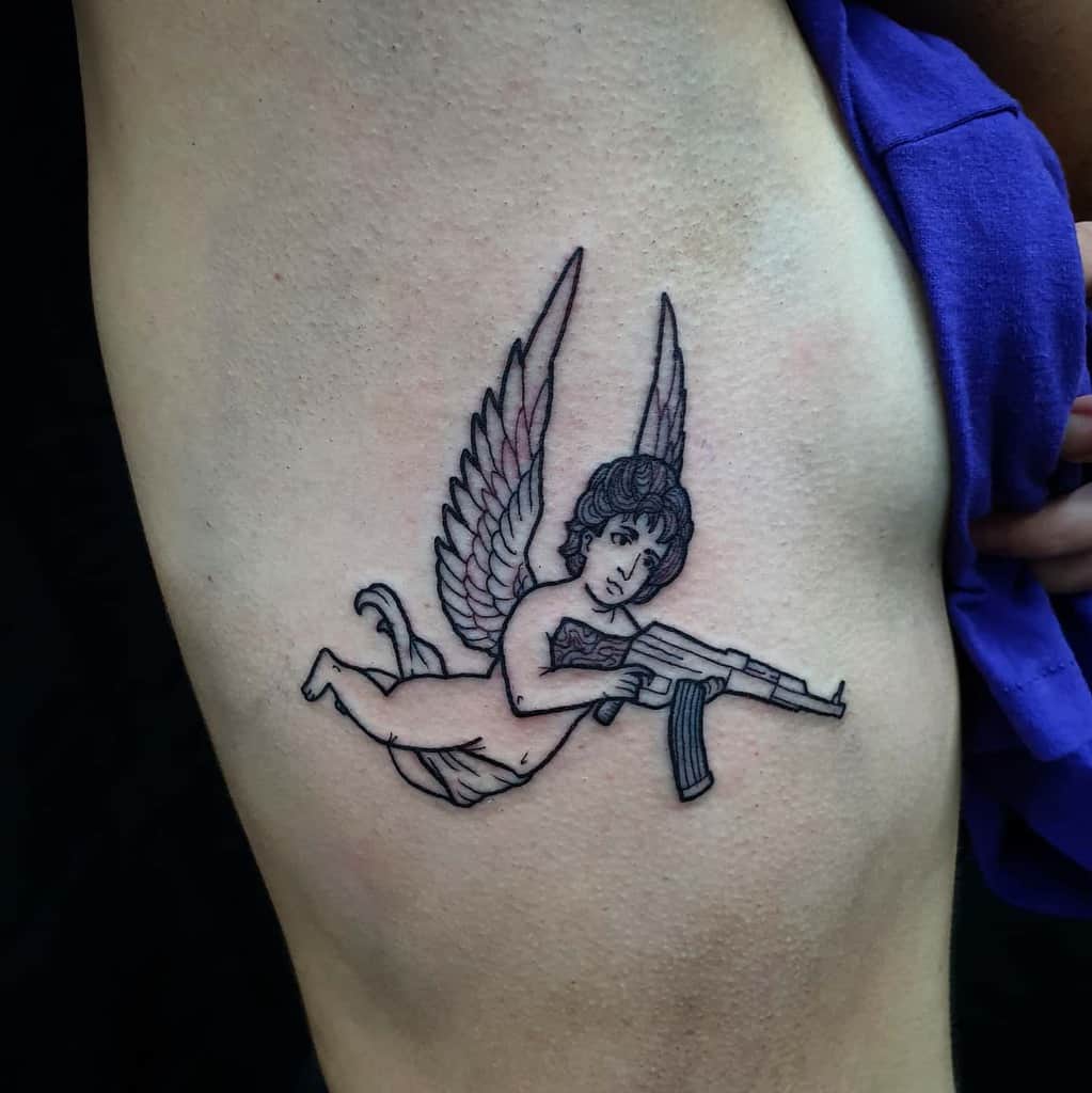 Tattoo of a cherub with wings, holding a rifle, on a person's ribcage