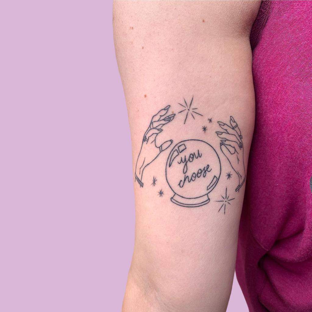 Tattoo of hands around a crystal ball with "you choose" text, surrounded by stars on an arm